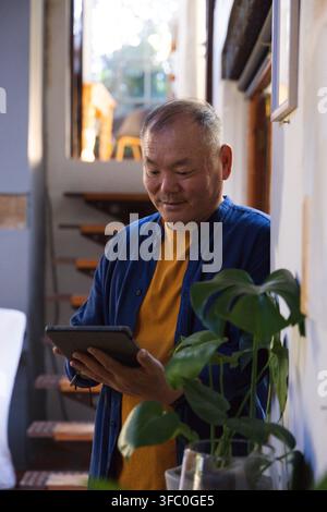 Seniorer asiatischer Mann steht in einem schmalen Flur und hält ein Tablet in der Nähe einer Holztreppe und einer Topfpflanze Stockfoto