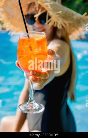 Frau mit Strohhut, die ein Glas Aperol-Spritz-Cocktail in der Nähe des Pools anbietet. Stockfoto
