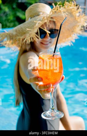 Frau mit Strohhut, die ein Glas Aperol-Spritz-Cocktail in der Nähe des Pools anbietet. Stockfoto