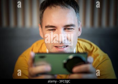 Glücklicher Mann mit Ohrstöpseln, der zu Hause Videos auf dem Smartphone anschaut. Junger Mann, der in Nahaufnahme auf seinem Telefon spielt Stockfoto