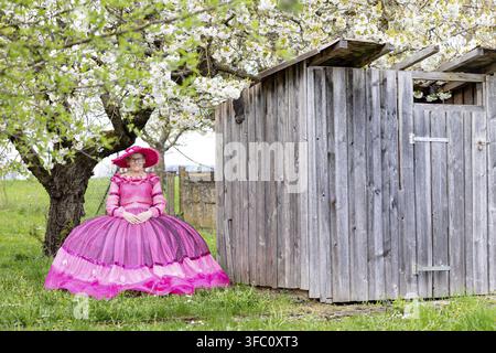 Frau in einem rosa Kleid, die unter blühenden Bäumen neben einem kleinen Holzschuppen in einer ländlichen Gegend sitzt, Marktoffingen, Donau-Ries-Kreis, Bayern, Deutschland Stockfoto
