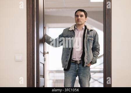 Junger Mann steht in einem Türrahmen, hält den Griff und ist dabei, einen Raum zu betreten. Beginn eines Arbeitstages, nach Hause kommen oder Einchecken in ein Hotel. Stockfoto