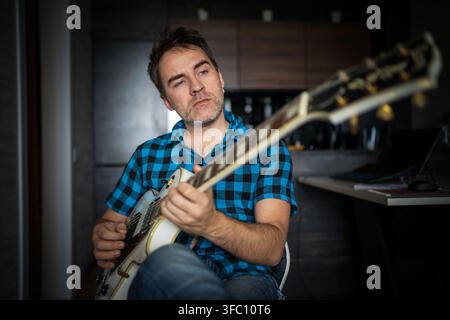 Mann spielt Gitarre, während er auf einem Stuhl sitzt, musikalisches Hobby und entspannte Atmosphäre zu Hause. Stockfoto