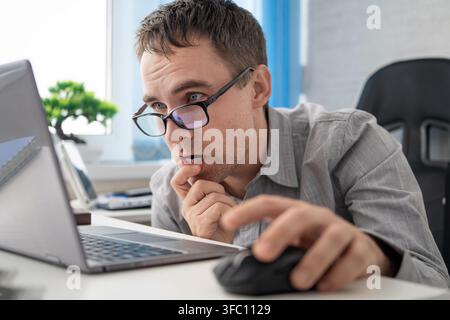 Ein überraschter Büroangestellter in einem grauen Hemd und einer Brille reagiert auf etwas auf dem Laptop-Bildschirm; er hält mit einer Hand eine Maus und berührt mit der anderen sein Kinn Stockfoto