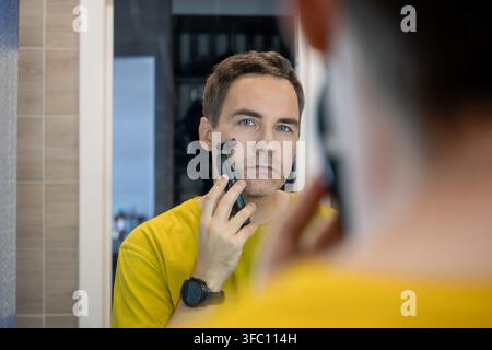 Mann rasiert sich mit einem Elektrorasierer vor dem Spiegel. Morgenpflegeroutine. Stockfoto