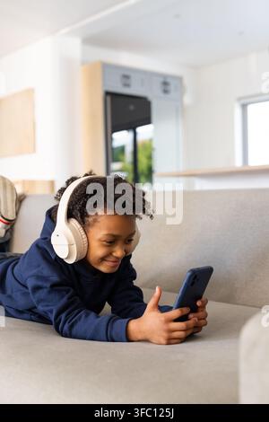Mädchen liegt auf grauem Sofa zu Hause, trägt marineblauen Hoodie, cremefarbene Kopfhörer und hält ein Smartphone Stockfoto