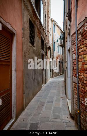 Schmale steingepflasterte Gasse zwischen alten Ziegel- und Putzgebäuden in Venedig, Italien, mit verwitterten Wänden, Fensterläden und ruhigem historischem Charme. Stockfoto