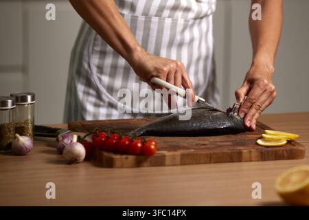 Frauenhände, die eine Schürze tragen, bereiten frischen Umberfisch auf einem Schneidebrett mit Kräutern, Kirschtomaten, Zitrone und Knoblauch zu Stockfoto