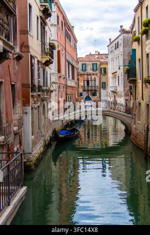 Schmaler Kanal in Venedig, Italien, gesäumt von verwitterten farbenfrohen Gebäuden, Blumenkästen, kleinen Booten und einer Stein-Fußgängerbrücke, die sich im ruhigen grünen Wasser spiegeln Stockfoto
