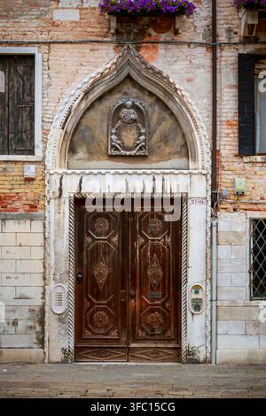 Verzierte hölzerne Doppeltür in einer verwitterten Ziegelmauer, eingerahmt von einem spitz zulaufenden Steinbogen und dekorativem Relief in einer historischen italienischen Straße. Stockfoto