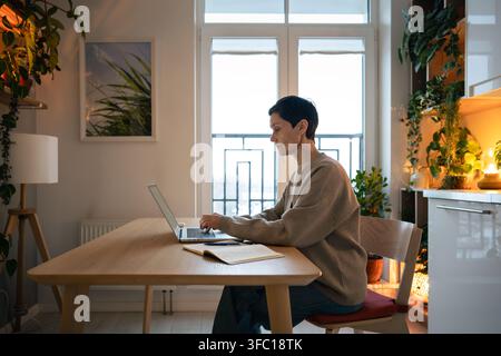 Reife Frau als Freiberuflerin arbeitet an einem Laptop in einem gemütlichen Wohninterieur mit Zimmerpflanzen und warmer Beleuchtung Stockfoto