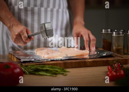 Koch mit Schürze bereitet rohe Hühnerfilets auf Aluminiumfolie mit einem Fleischklopfer in einer hellen Kücheneinrichtung zu Stockfoto
