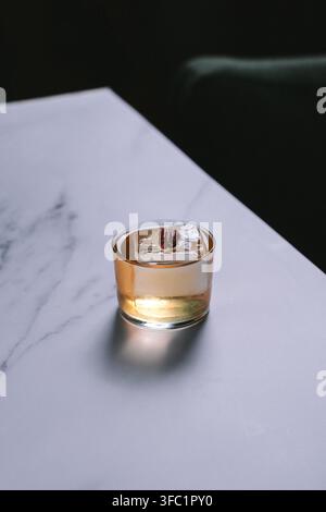 Blasser goldener Cocktail auf einem großen Eiswürfel in einem Rocks-Glas an einer weißen Marmortisch-Ecke vor dunklem Hintergrund, minimalistische Getränkefoto Stockfoto