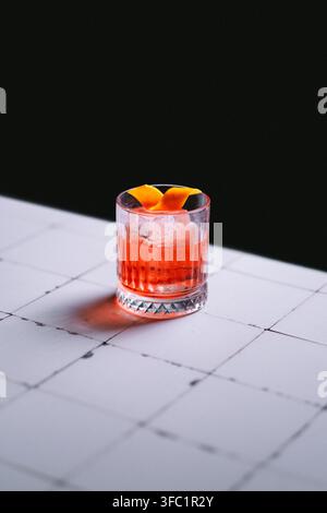 Negroni on the rocks in einem gerippten Rocks-Glas mit einer Orangenschale auf weißen Fliesen vor einem schwarzen Hintergrund, minimalistische stimmungsvolle Getränke Stockfoto