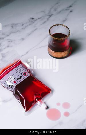 Halloween-Blutbeutel-Cocktail mit einem gewürzten Rand im Tumbler aus rotem Getränk und roten Tropfen auf einem weißen Marmortisch, gruselig thematisierte Partygetränke Stockfoto