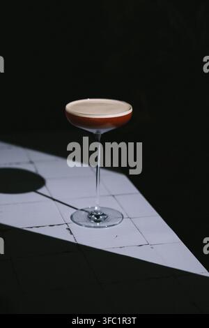 Dunkler roter Coupe-Cocktail mit cremigem Schaum auf einem sonnenbeschienenen weißen Fliesentisch mit einem langen Schatten vor schwarzem, minimalistischem, stimmungsvoller Getränkefoto Stockfoto