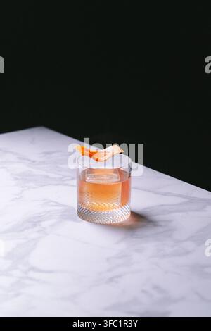 Orangener Old Fashioned in einem Kristall-Rocks-Glas mit einer getrockneten Orangenzange auf weißem Marmor vor schwarzem Hintergrund, elegante stimmungsvolle Getränkefotografie Stockfoto