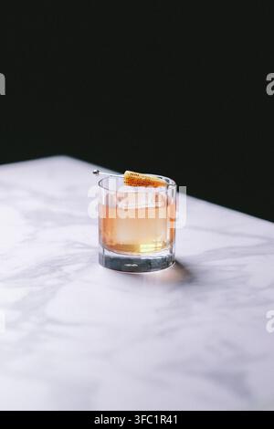 Amber-Cocktail auf Eis in einem geschliffenen Kristalltumbler mit Orangenschale auf weißem Marmor vor schwarzem Hintergrund, elegante stimmungsvolle Getränkefotografie Stockfoto