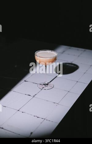 Coupe-Cocktail mit hellrosa Schaum auf einem weiß gekachelten Tisch in einem harten Lichtstrahl vor schwarzem Hintergrund, langer Schatten, minimalistische stimmungsvolle Getränkefotografie Stockfoto