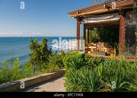 Eine malerische Außenterrasse eines Restaurants, umgeben von üppiger Vegetation, bietet einen malerischen, erhöhten Blick auf einen sandigen Strand und blaue Meereswellen unter einem klaren Stockfoto