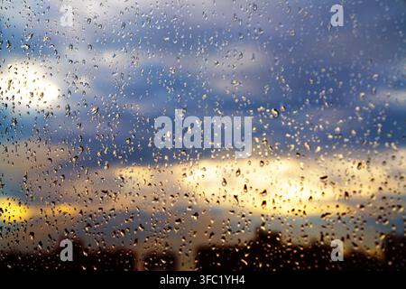 Eine stimmungsvolle Ansicht einer verschwommenen Stadtsilhouette durch ein regennasses Fenster, mit Wassertropfen auf dem Glas vor einem bewölkten Himmel. Stockfoto