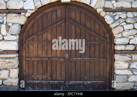 Eine robuste, gewölbte Holztür mit kunstvollem Eisenschmuck und Ringgriffen, eingebaut in eine rustikale Steinmauer. Stockfoto