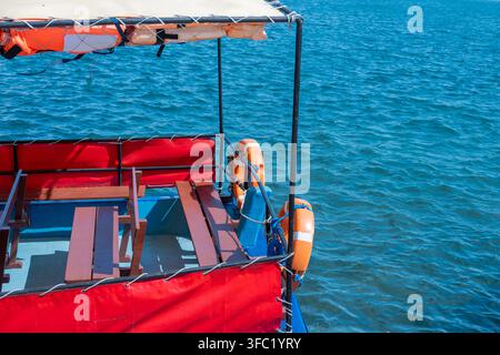 Ein leeres blaues Passagierboot mit Holzbanken und einem Sonnensegel, das mit Schwimmwesten gesichert ist, schwebt auf ruhigem blauem Wasser mit einem belebten Hafen im Stockfoto