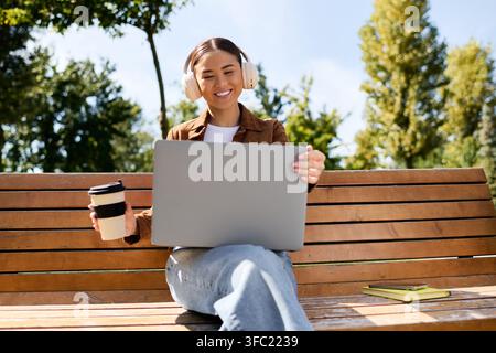 Eine fröhliche junge Frau lächelt auf einer Bank, nippt an ihrem Kaffee und nutzt ihren Laptop zwischen Bäumen. Stockfoto