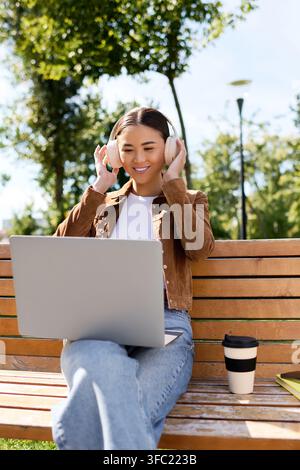 Lächelnde junge Frau mit Kopfhörern arbeitet an ihrem Laptop, umgeben von Natur in einem Park. Stockfoto