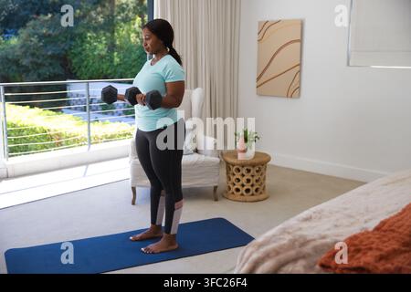 Afroamerikanische Frau trainiert auf blauer Matte in einem sonnigen Schlafzimmer am Balkon und hält schwarze Sechskantgewichte Stockfoto