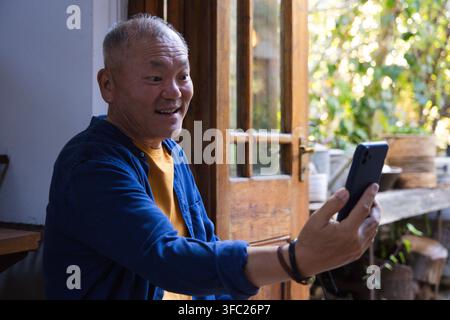 Seniorer asiatischer Mann in blauem Hemd hält ein schwarzes Telefon und schaut auf einen Bildschirm in der Nähe einer offenen Holztür Stockfoto