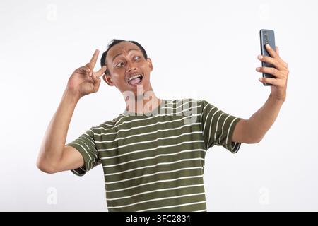 Glücklicher junger Asiate in einem grünen Hemd lacht, während er sein Smartphone hält und eine Friedensgeste mit dem Finger zeigt, isoliert auf weißem Hintergrund. Stockfoto