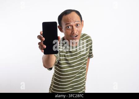 Ein asiatischer Mann in einem grünen Hemd lächelt, während er ein Smartphone mit einem leeren Bildschirm hochhält. Der Hintergrund ist einfarbig weiß, und der Mann zeigt auf die Ka Stockfoto