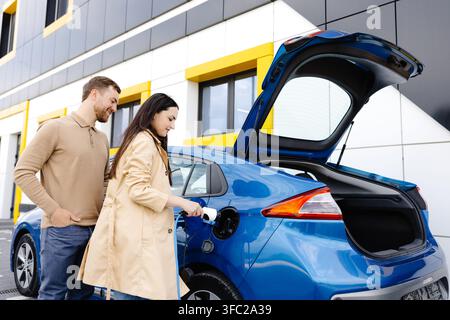 Ein junges Paar, das mit einem Elektroauto unterwegs ist und an der Ladestation hält. Eine Freundin, die das Ladegerät hält, das Auto ansieht und lächelt. Stockfoto