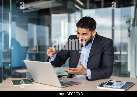 Gespannter Geschäftsmann im Anzug, der während einer emotionalen Online-Videokonferenz in einem modernen Büro schreit und auf den Laptop-Bildschirm zeigt, Frustration und Kommunikationsprobleme erlebt Stockfoto