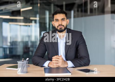 Selbstbewusster junger indischer Geschäftsmann sitzt an einem Bürotisch mit verschränkten Händen und verkörpert Professionalität, Führung und Erfolg in einem modernen Unternehmensklima Stockfoto
