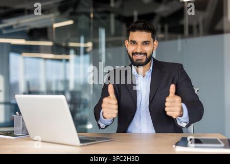 Junger männlicher Profi zeigt Daumen-hoch-Gesten und lächelt in die Kamera, was Zustimmung, Erfolg und positives Feedback in einem modernen Büroumfeld darstellt Stockfoto