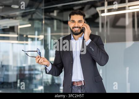 Selbstbewusster indischer Geschäftsmann, der in einem modernen Firmenbüro am Mobiltelefon spricht, Brillengläser hält und lächelt, repräsentierend Kommunikation, Erfolg und berufliches Networking Stockfoto