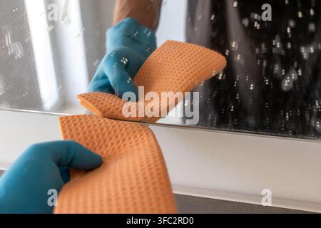 Nahaufnahme von Händen in blauen Schutzhandschuhen, die eine verschmierte Spiegeloberfläche mit einem strukturierten orangefarbenen Reinigungstuch abwischen Stockfoto