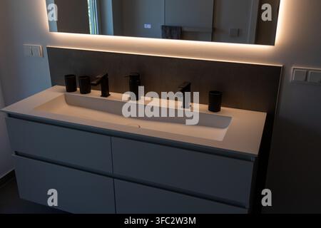 Ein schlankes, zeitgenössisches Badezimmerwaschtisch verfügt über ein weißes integriertes Waschbecken mit zwei schwarzen Wasserhähnen und umgebender LED-Hintergrundbeleuchtung Stockfoto