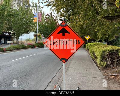 Hellorangefarbenes rautenförmiges Baustellenschild mit einem Pfeil, das auf einem Gehweg neben einer städtischen Straße angebracht ist Stockfoto
