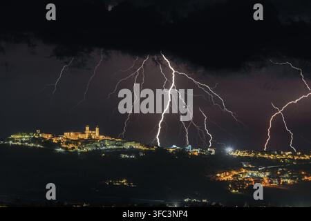 Blick auf Blitze, die den Nachthimmel über der historischen Hügelstadt Montecosaro mit leuchtenden Straßenlaternen und dunklen Gewitterwolken erhellen, Mo Stockfoto
