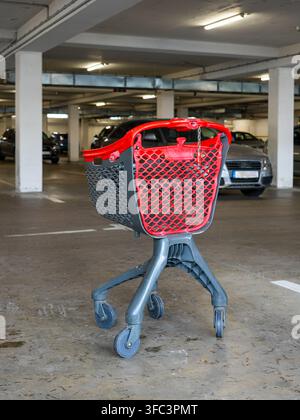 Leerer roter und grauer Einkaufswagen steht allein in einer unterirdischen Parkgarage mit geparkten Autos und repräsentiert Einzelhandel, Einkaufen und Verbraucherlebensstil Stockfoto