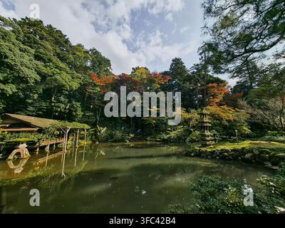 Blick auf einen ruhigen Teich im Kenrokuen-Garten mit einem traditionellen hölzernen Teehaus, einer Steinlaterne und lebhaftem Herbstlaub in Kanazawa, Ishikawa, Japan. Stockfoto