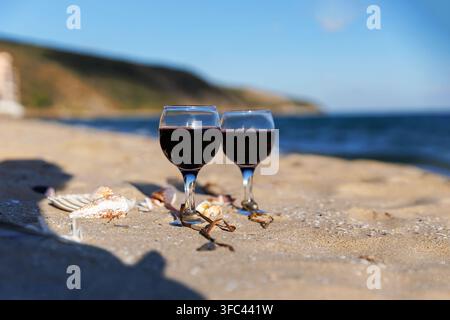 Zwei Gläser Rotwein auf sandigem Strand mit Meeresmuscheln, Ozeanwellen und Küstenklippen, malerische Sommer-Meeresentspannung Stockfoto