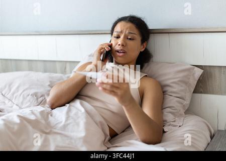 Junge schwarze Frau, die am Smartphone spricht, während sie ein Thermometer im Bett überprüft, Rat oder Unterstützung sucht, während sie sich krank fühlt und ihre Gesundheit überwacht Stockfoto