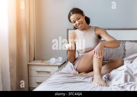 Schwarze Frau lächelt, während sie Feuchtigkeitscreme auf ihr Bein im Bett aufträgt Stockfoto