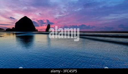 Blick auf Haystack Rock bei Sonnenuntergang mit lebhaften violetten und rosa Wolken, die sich im nassen Sand des Strandes in Cannon Beach, Oregon, USA, spiegeln Stockfoto
