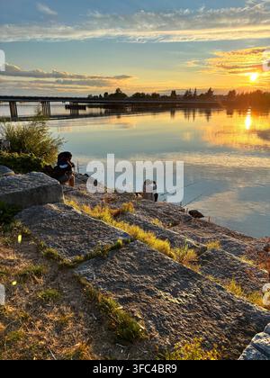 Zwei Menschen fischen von einer felsigen Küste bei Sonnenuntergang, ihre Ruten schweben über ruhigem Wasser, das im goldenen Licht glüht Stockfoto
