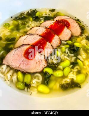 In Scheiben geschnittenes Schweinefilet, serviert über Nudeln, Edamame und Grünkohl in einer klaren Brühe mit scharfer roter Currysauce. Stockfoto